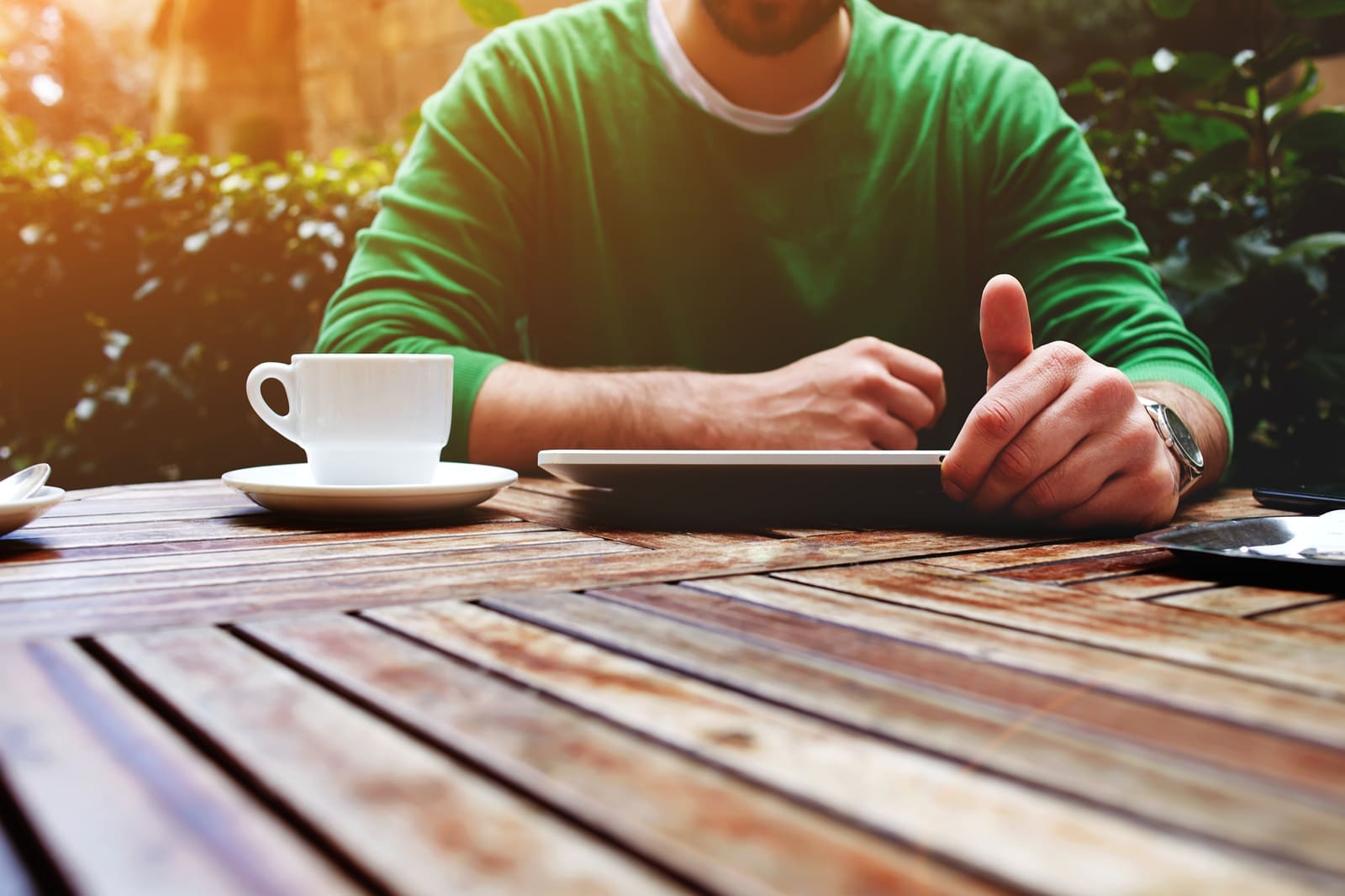 Young man sitting at the table with cup of coffee digital tablet Front view young man sitting at the table with cup of coffee digital tablet and smart phone man's hands texting message on touch screen tablet while sitting on balcony terrace with plants flare